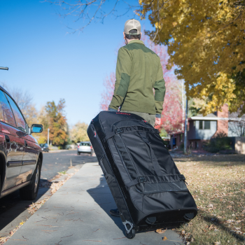SKB-Nylontasche mit Rollen, von einer Person auf einem Bürgersteig in einem Wohngebiet mit geparkten Autos und Herbstbäumen gezogen.